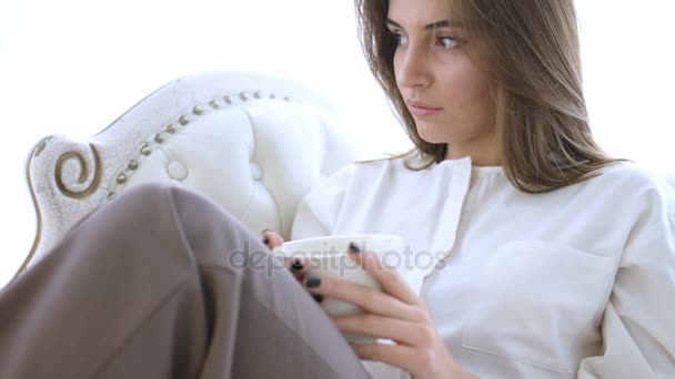 Mulher bonita sonhando com xícara de café quente ou chá sentado no sofá — Vídeo de Stock