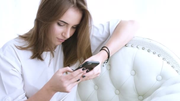美しい少女はソファーに座って携帯電話でテキスト メッセージ。女性のスマート フォンのアプリを使って — ストック動画