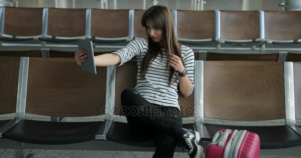 Jovem garota tirando uma selfie sentada na cadeira no aeroporto Jovem morena tira foto de si mesma no tablet pc, uma selfie — Vídeo de Stock