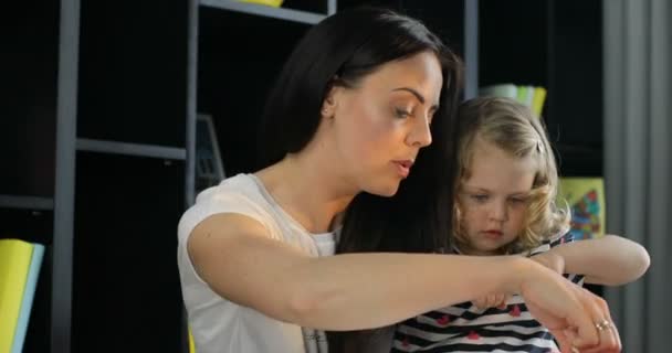 Jovem caucasiana com sua filhinha com cabelo encaracolado loiro desenhando em uma folha de papel sentada em uma mesa em um quarto. Interior . — Vídeo de Stock