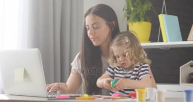 Güzel küçük kız sarışın kıvırcık saçlı annesi evde dizüstü bilgisayarda çalışma sırasında hamuru ile kutusunu açma. Kapalı.