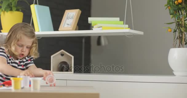 Kaukasische mooi meisje met blond krullend haar doos met plasticine terwijl haar moeder met behulp van laptop thuis. Indoor. — Stockvideo