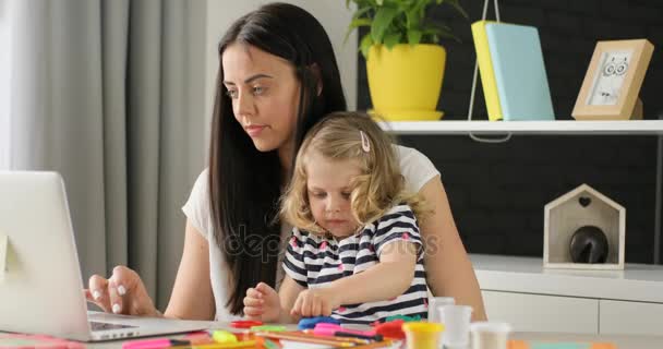 Caucasian young woman with black long hair using laptop for shopping while her dauther playing with plasticine on home background. — Stock Video