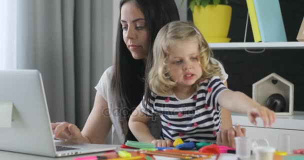 Mulher branca com cabelos longos pretos usando laptop para trabalhar enquanto sua filha esculpir a partir de plasticina no fundo de casa . — Vídeo de Stock