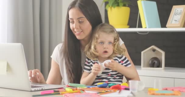 Carming holčička s blond kudrnaté vlasy modelování z plastelíny, zatímco její matka pomocí přenosného počítače na domácí pozadí. — Stock video