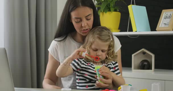 Charmig liten tjej med blont lockigt hår placering på skrivbordet i hem med sin mor och att göra bubble blower. Inomhus. — Stockvideo