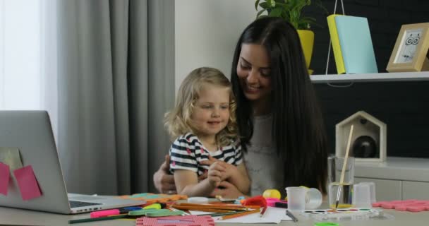 Menina branca encantadora com cabelo encaracolado loiro e sua mãe com cabelos longos pretos sentados na mesa no quarto. Interior . — Vídeo de Stock