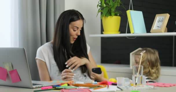 Mooie vrouw met lange zwarte haren aanbrengen op de tafel in de kamer terwijl haar weinig dauther met blond krullend haar, waardoor haar moeder de mobiele telefoon. Indoor. — Stockvideo