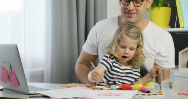 Beau jeune homme avec des lunettes et sa dauther avec des cheveux bouclés blonds dessin sur une feuille de papier en utilisant des peintures sur fond de maison . — Video