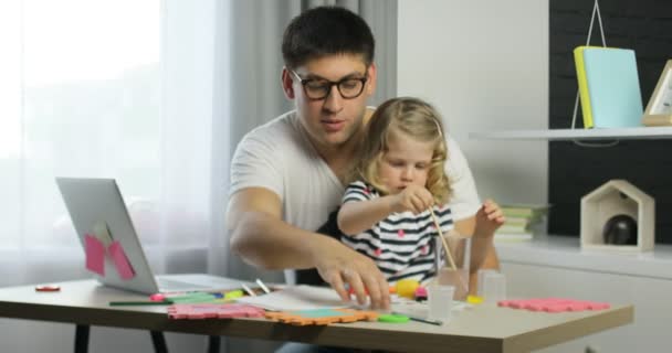 Caucasian little girl with blond curly hair using paints for drawing on a sheet of paper siting with her father in home. — Stock Video