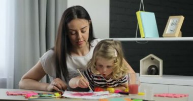 Beyaz anne ve küçük dauther sarışın kıvırcık saçlı boyalar için driwing kullanarak, kameraya bakıyor ve gülümsüyor. Kapalı.