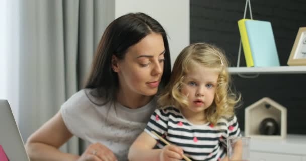 Aantrekkelijk meisje met blond krullend haar met haar moeder met behulp van verven voor driwing, op zoek op camera en glimlachen. Indoor. — Stockvideo