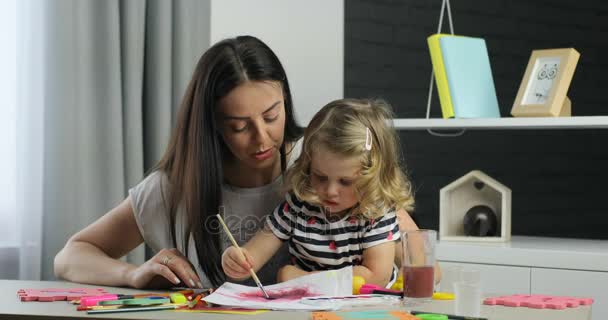 Donna caucasica con lunghi capelli neri e la sua piccola dauther con i capelli biondi ricci che gocciolano usando vernici, guardando sulla macchina fotografica e sorridendo con i denti. Interni . — Video Stock