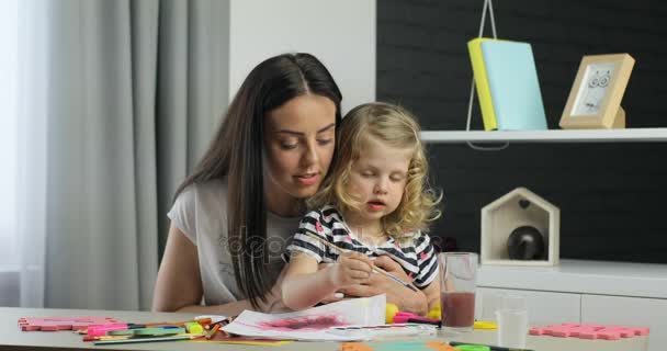 Vonzó fiatal nő, hosszú, fekete haja és ő kis dauther szőke göndör haja elhelyezés otthonában és használ festékek, driwing keresi a kamera, és mosolygott. Beltéri. — Stock videók