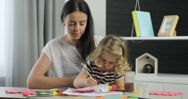 Encantadora niña con el pelo rubio rizado y su madre con el pelo largo y negro chorreando con pinturas, mirando a la cámara y sonriendo con los dientes. De interior . — Vídeos de Stock