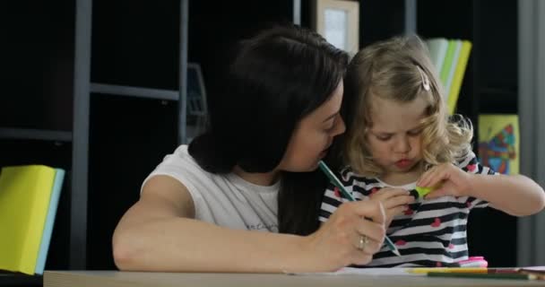 Attraente bambina con i capelli biondi ricci che disegna con sua madre da pennarello posizionato sul tavolo in casa. Interni . — Video Stock
