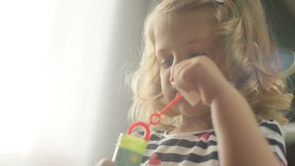 Portret van zijaanzicht van vrij Kaukasische jong meisje met blond krullend haar zeepbel blower op levende achtergrond maken. Indoor. — Stockvideo