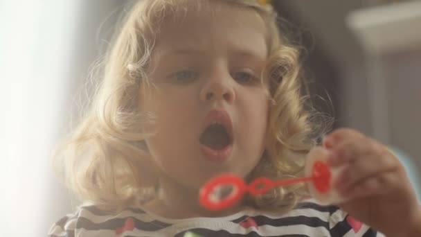 Portret van aantrekkelijke Kaukasische jong meisje met blond krullend haar en blauwe ogen zeepbel blower op home achtergrond maken. Indoor. — Stockvideo