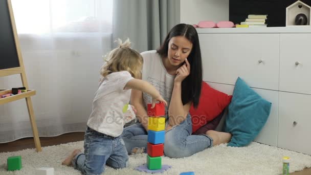 Charmind liten flicka med blont lockigt hår och hennes mor med svart långt hår placering på golvet i rummet och leker med kuber. Inomhus. — Stockvideo
