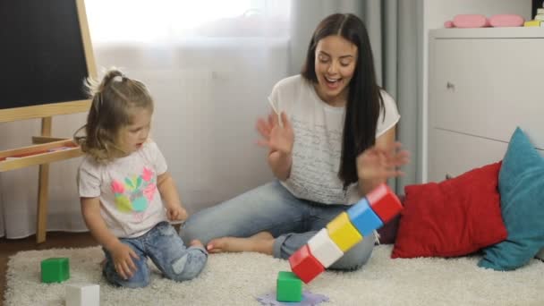 Bella bambina con i capelli biondi ricci e sua madre con i capelli lunghi neri che gioca con i cubi situati sul pavimento in camera. Interni . — Video Stock