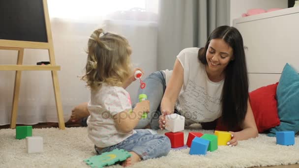 Ragazzina caucasica carina con i capelli biondi ricci che si siede sul pavimento in casa con la madre e gioca con i cubi. Interni . — Video Stock