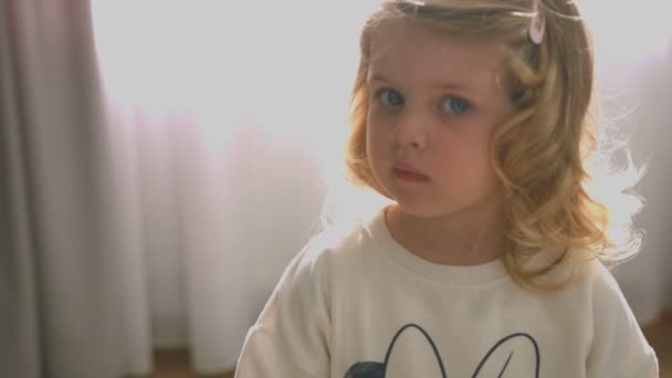 Porträt eines ernsthaften kaukasischen Mädchens mit blonden lockigen Haaren und blauen Augen, das vor heimischem Hintergrund in die Kamera blickt. Innenräume. — Stockvideo