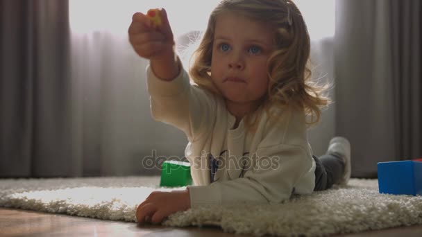 Carmind niña con el pelo rubio rizado acostado en el suelo y jugando el pequeño juguete en el fondo de la casa. De interior . — Vídeos de Stock