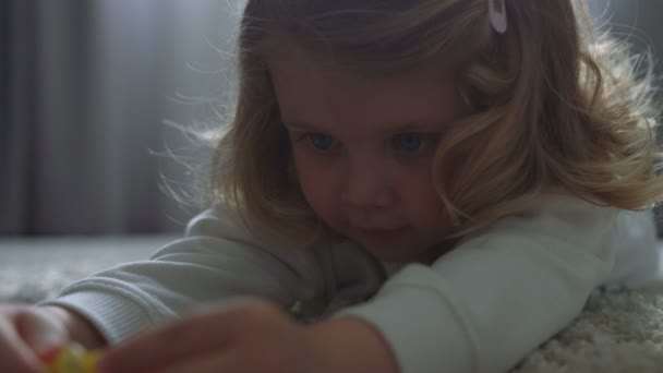 Retrato de menina bonita com cabelo encaracolado loiro e olhos azuis brincando de brinquedo deitado no chão em casa. Interior . — Vídeo de Stock