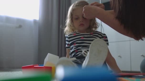 Ragazzina attraente caucasica con i capelli biondi ricci che si trova sul pavimento su sfondo vivente. Mamma che aggiusta i peli di Dauther. Interni . — Video Stock