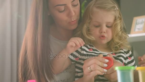 Giovane donna caucasica con lunghi capelli neri e il suo charmind piccolo dauther con i capelli biondi ricci aprendo la scatola di plastilina su sfondo vivente. Interni . — Video Stock