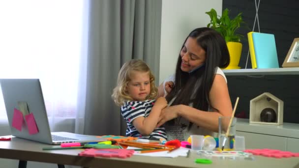 Jonge vrouw met lange zwarte haren aanbrengen op het bureau thuis met haar kleine dauther met blond krullend haar en praten over de mobiele telefoon. Indoor. — Stockvideo