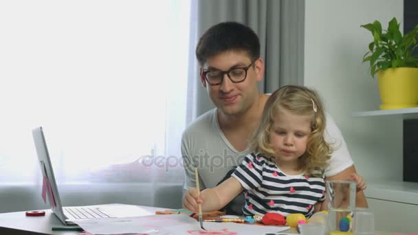 Charmantes kleines Mädchen mit blonden lockigen Haaren, während ihr Vater mit roter Farbe und Pinsel auf Papier malte. Innenräume. — Stockvideo