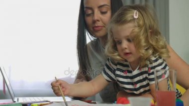 Sevimli küçük kız sarışın kıvırcık saçlı'ana tabloda siting ve annesi boya ve boya fırçası kullanarak resim. Kapalı.