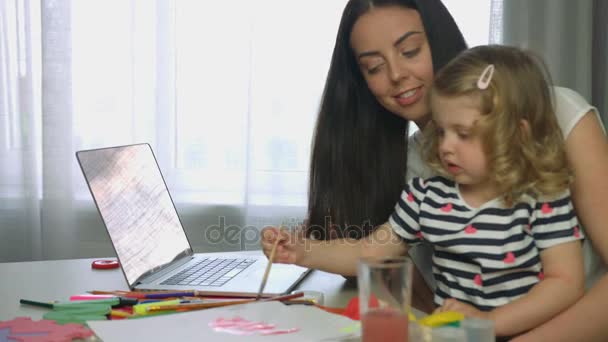 Zijaanzicht van meisje met blond krullend haar schilderen met verf en penseel op vel papier en haar moeder plaatsing op het Bureau. Indoor. — Stockvideo