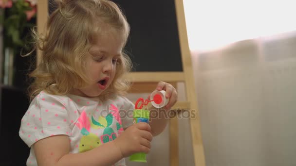 Menina bonita caucasiana com cabelo encaracolado loiro fazendo soprador de bolhas no fundo da casa. Interior . — Vídeo de Stock