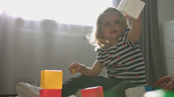 Vista lateral de charmind caucásica niña con el pelo rizado rubio sentado en el suelo en casa y jugando con cubos y juguetito. De interior . — Vídeo de stock