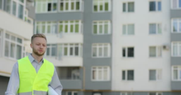 Beau constructeur caucasien dans un gilet vert allant sur fond de construction inachevé mettant sur le casque blanc, regardant autour. Extérieur . — Video