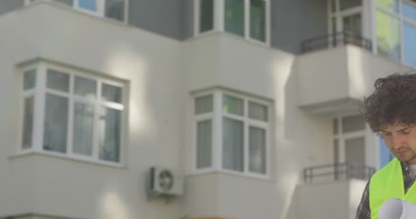 Портрет молодого кавказского строителя с черными вьющимися волосами в зеленом жилете, стоящего на незавершенном строительном фоне, надевающего желтый шлем, смотрящего в сторону. На открытом воздухе . — стоковое видео
