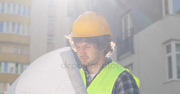 Arquitecto caucásico en casco amarillo sobre fondo de edificio. Ingeniero en un sitio de construcción buscando dibujo. Exterior . — Vídeos de Stock