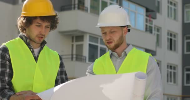 Двоє чоловіків-будівельників у захисному шоломі на голові та спеціальному одязі дивляться малюнок будівлі на незакінченому будівництві. На відкритому повітрі . — стокове відео