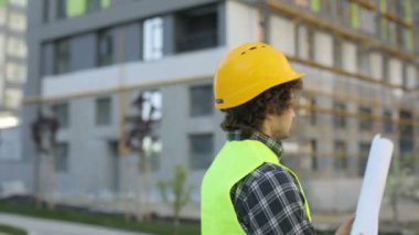 Bina arka plan üzerinde duran sarı kask içinde tutarak ve çizim dağıtma genç mimar görünümünü geri. Açık.