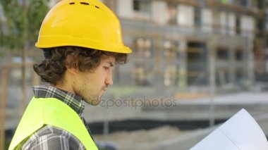 Yan portresi ciddi Builder sarı kask ve bitmemiş inşaat toprakları üzerinde duran, çizim ve bina bakarak yeşil yelek görüntüleyin. Açık.