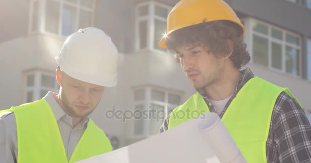 Retrato de dois construtores mais jovens em um capacete protetor em suas cabeças e roupas especiais olhando desenho do edifício e falando sobre isso. Ao ar livre . — Vídeo de Stock