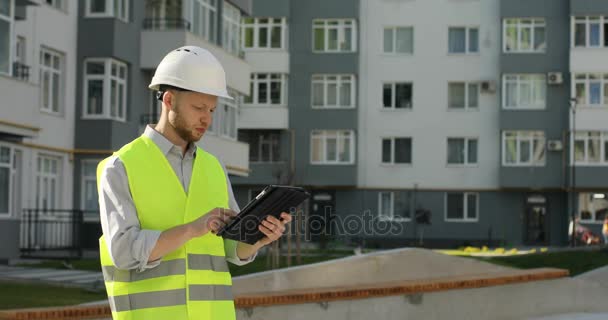 彼の頭と未完成の建物が背景の彼の仕事のためのタブレットを使用して緑色のベストに白いヘルメットを保護で若いビルダーが建築家します。. — ストック動画