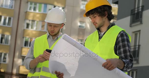 Kaukasischer Bauarbeiter mit weißem Helm, der auf dem Handy telefoniert, während sein Kollege Zeichnungen auf dem unfertigen Bauhintergrund hält. Nahaufnahme. — Stockvideo