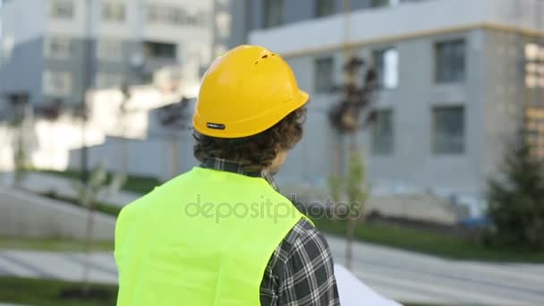 Vista posterior del arquitecto en casco amarillo y chaleco verde de pie sobre el fondo del edificio, analizando el dibujo. Exterior . — Vídeo de stock
