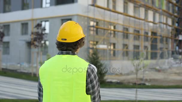 Vista posteriore di architetto caucasico in casco giallo e giubbotto verde cercando disegno in piedi su sfondo edificio. All'aperto . — Video Stock