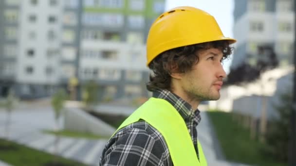 Retrato de vista lateral do construtor concentrado caucasiano em capacete amarelo e colete verde em pé sobre fundo de construção inacabado . — Vídeo de Stock