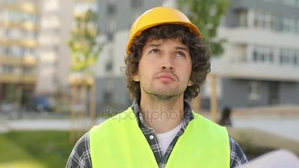 Портрет молодого архітектора в жовтому шоломі і зеленому жилеті, що стоїть на незакінченій будівельній території, дивлячись вгору і на малюнок. На відкритому повітрі . — стокове відео