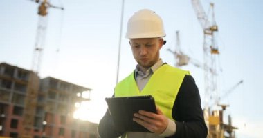 Beyaz kask ve yeşil yelek bitmemiş inşaat arka plan üzerinde çalışmak için tablet kullanarak ciddi Oluşturucu.